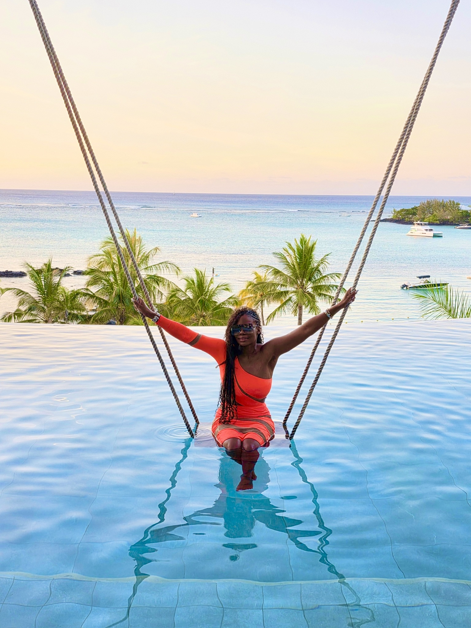 Jazzee smiling on a swing over an infinity pool overlooking the ocean and palm trees at sunset